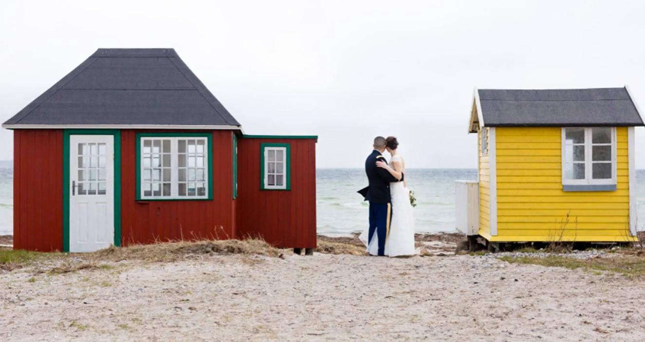 A couple introduced by Annabel, standing together looking out to sea between two beach huts